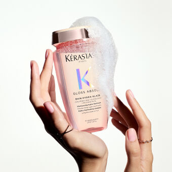 Flacon de shampoing Kerastase Gloss Absolu, rempli de mousse, tenu par des mains delicates sur fond clair.