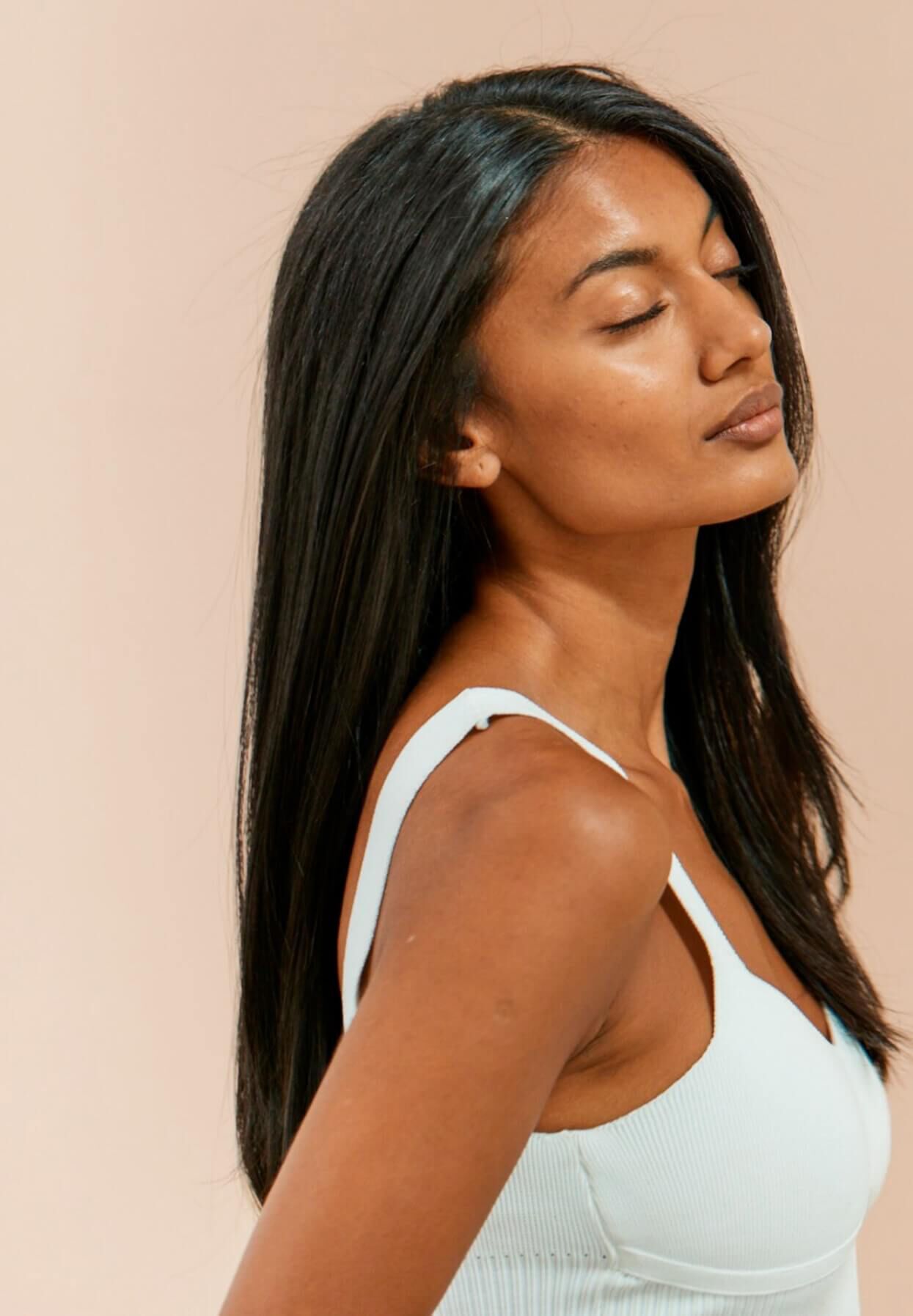 A woman with long, straight black hair looks serene, with her eyes closed and head slightly tilted against a soft, neutral background. She is wearing a light blue tank top.