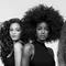 A black-and-white portrait of three women standing side by side, each showcasing different hairstyles: one with loose curls, one with a large afro, and one with wavy red hair. They are wearing sleeveless black tops and are posed against a plain background.
