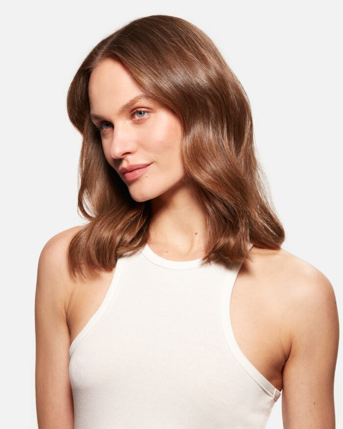 Femme avec des cheveux bruns ondules, portant un top sans manches blanc, regardant legerement sur le cote.