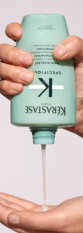 A person's hand is holding a turquoise Kerastase hair product bottle, pouring a creamy substance into the other hand.