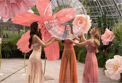 Trois femmes en robes fluides interagissant avec des bulles transparentes et des fleurs geantes roses dans un cadre luxuriant.