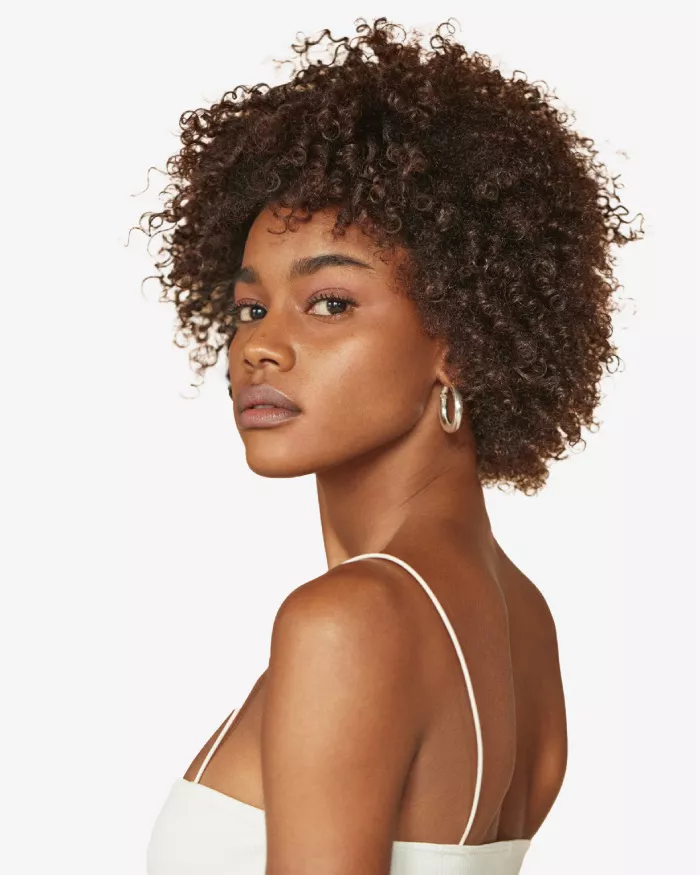 Portrait d'une femme aux cheveux boucles, regardant par-dessus son epaule, avec un fond blanc. Elle porte un debardeur et des boucles d'oreilles.