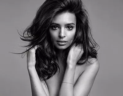 A black and white portrait of a woman with long, flowing hair, posing with her hands gently touching her arms and looking directly at the camera.