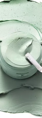 A jar of green hair mask surrounded by a smooth, textured application of the same product, with a metal spatula resting on top.