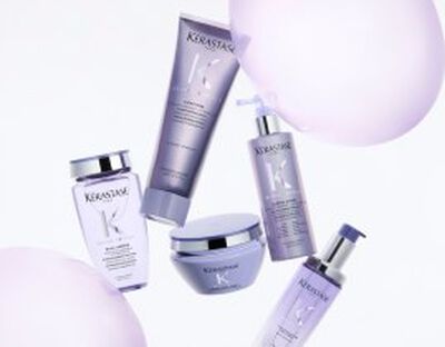 A collection of Kerastase hair care products displayed elegantly on a iridescent background. The products include a shampoo, conditioner, a treatment, and a serum, all presented in sleek packaging with a lavender theme.