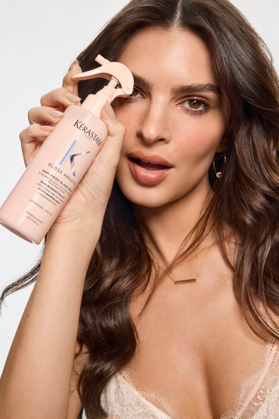 Une femme posant avec un flacon de produit capillaire Kerastase, mettant en valeur ses cheveux ondules et son maquillage naturel.