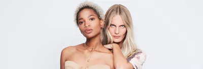 Two women pose closely together against a plain white background. One has curly blonde hair and is wearing a strapless top, while the other has long straight hair and a patterned shirt. They both have confident expressions.