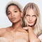 Two women pose closely together against a plain white background. One has curly blonde hair and is wearing a strapless top, while the other has long straight hair and a patterned shirt. They both have confident expressions.