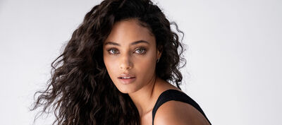 A young woman with long, wavy dark hair and striking green eyes poses against a light gray background, wearing a black sleeveless top.