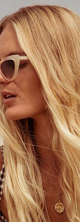 A woman with long, wavy blonde hair and cat-eye sunglasses, gazing thoughtfully into the distance against a sandy backdrop.