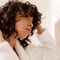 A person with curly hair adjusts their hair while wearing a white bathrobe, standing in a softly lit bathroom.