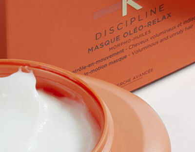 A close-up of a creamy white hair mask in an orange jar, with a partially open lid, next to an orange box labeled "Discipline Masque Oleo-Relax."