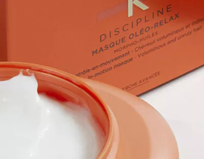 A close-up of a creamy white hair mask in an orange jar, with a partially open lid, next to an orange box labeled "Discipline Masque Oleo-Relax."