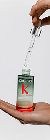 A hand holding a bottle of Kerastase Genesis Homme serum, with a dropper releasing a drop of serum above it. The bottle has a gradient color from green to orange.