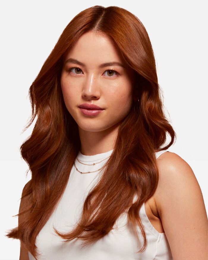 Une femme avec des cheveux longs et roux, portant un haut blanc, regarde l'objectif avec un leger sourire.