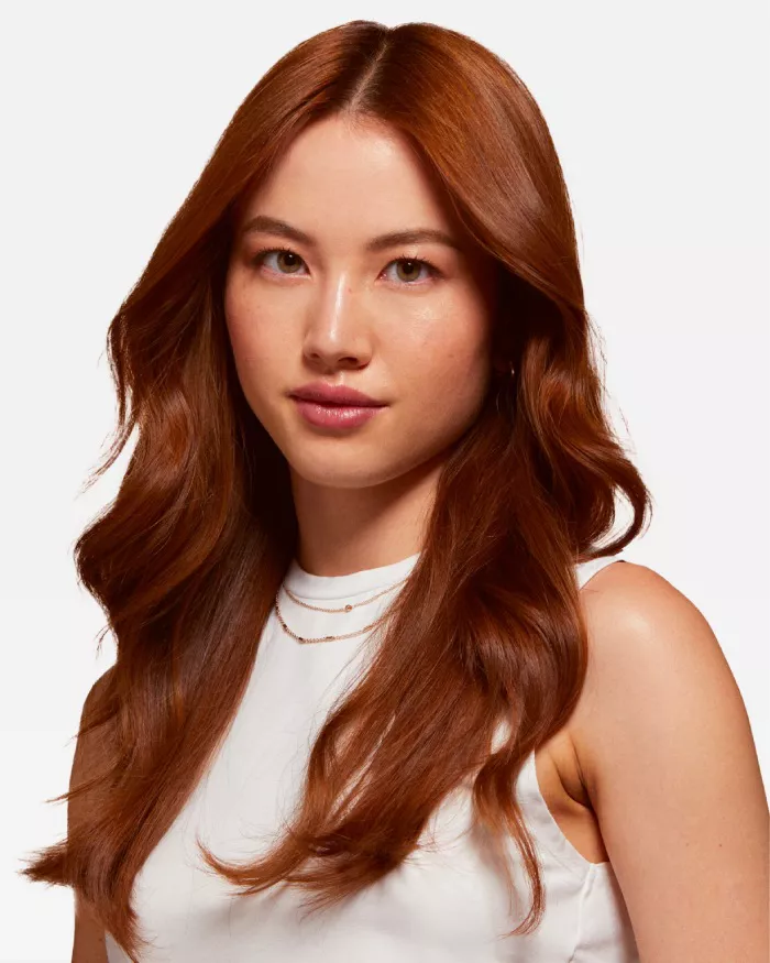 Une femme avec des cheveux longs et roux, portant un haut blanc, regarde l'objectif avec un leger sourire.