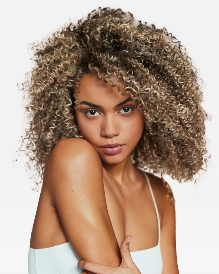 Portrait d'une femme aux cheveux boucles et blonds, avec un regard expressif. Elle pose en tenant son bras, sur un fond neutre.
