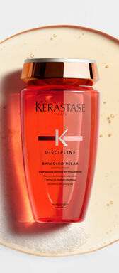 A bottle of Kerastase Discipline Bain Oleo-Relax shampoo, featuring a sleek design in a vibrant orange hue, set against a circular splash of water.
