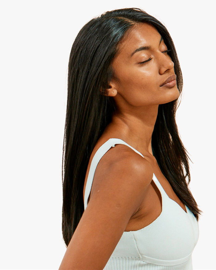 Une femme d'origine indefinie avec de longs cheveux noirs, les yeux fermes et une expression sereine, porte un debardeur blanc et se tient legerement de profil.