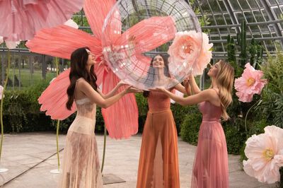Three women in a botanical setting are joyfully interacting with a large, transparent orb surrounded by vibrant flower decorations.