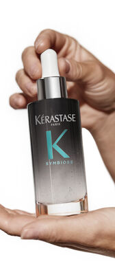 A hand holding a bottle of Kerastase Symbiose hair serum, featuring a sleek design with a gray gradient and a dropper top.