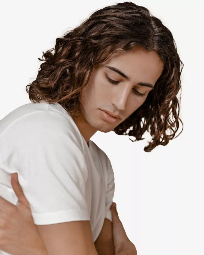 Un jeune homme aux cheveux boucles, portant un t-shirt blanc, se tourne legerement sur le cote, avec un regard pensif et une expression contemplative.