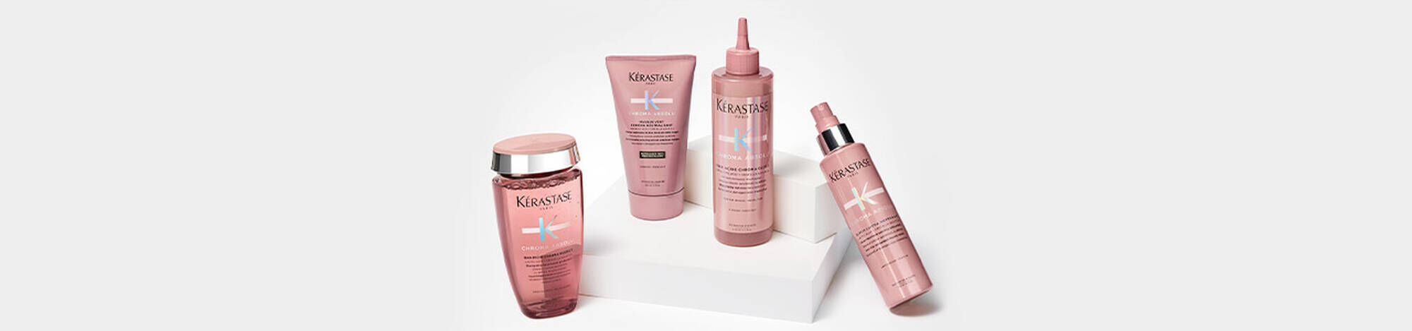 Image of a collection of hair care products displayed on a white pedestal. The products, all in pink packaging, include a shampoo, a hair mask, a leave-in treatment, and a spray.