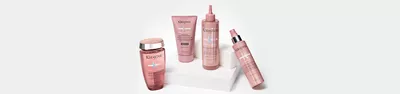 Image of a collection of hair care products displayed on a white pedestal. The products, all in pink packaging, include a shampoo, a hair mask, a leave-in treatment, and a spray.