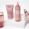 Image of a collection of hair care products displayed on a white pedestal. The products, all in pink packaging, include a shampoo, a hair mask, a leave-in treatment, and a spray.