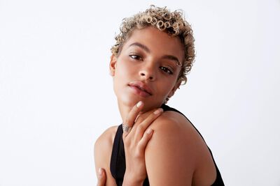 A close-up portrait of a person with curly hair, looking thoughtfully at the camera, with a soft expression against a plain background.
