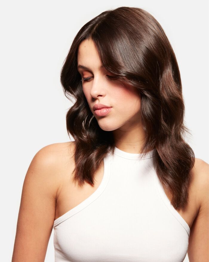 Femme avec des cheveux bruns ondules, regardant vers le bas, portant un haut sans manches blanc.