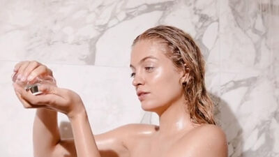 A person with wet hair holds a small jar in one hand, looking thoughtfully at the contents, in a marble bathroom setting.