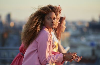 A woman with long, curly hair gazes thoughtfully into the distance, while two other individuals are seen in profile beside her against a sunset backdrop.