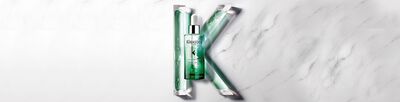 A green bottle of hair serum is displayed at the center, surrounded by flowing green gel tubes on a white marble background, with a stylized letter "K" formed by the arrangement.
