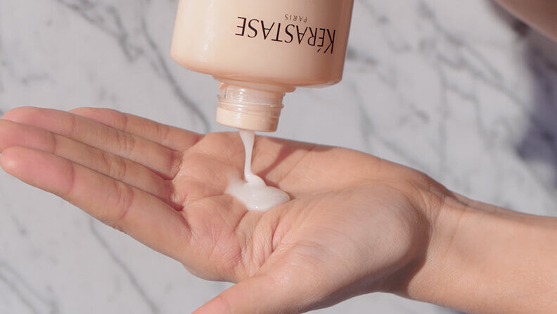 A person's hand holding a bottle of Kerastase hair product, with a small amount of creamy lotion being poured into the palm. The background features a marble surface.