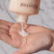 A person's hand holding a bottle of Kerastase hair product, with a small amount of creamy lotion being poured into the palm. The background features a marble surface.