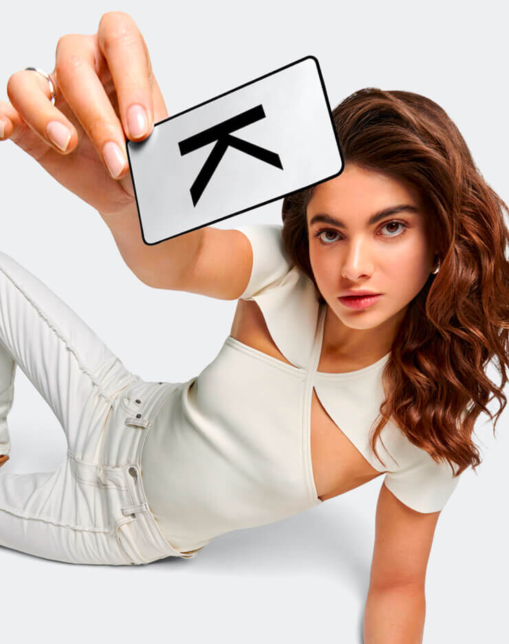 Une jeune femme aux cheveux bruns fonces, vetue d'un haut creme a decoupes et d'un pantalon clair, est allongee sur un fond gris clair, regardant directement l'objectif. Elle tient une carte de fidelite Kerastase dans sa main.