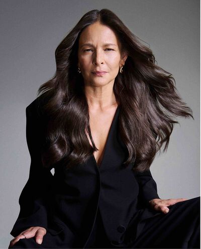 Portrait d’une femme mure aux longs cheveux bruns volumineux portant un blazer noir sur un fond gris.
