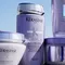 Four Kerastase hair care products in purple containers are displayed against a bright background, featuring labels with the brand name and product details.