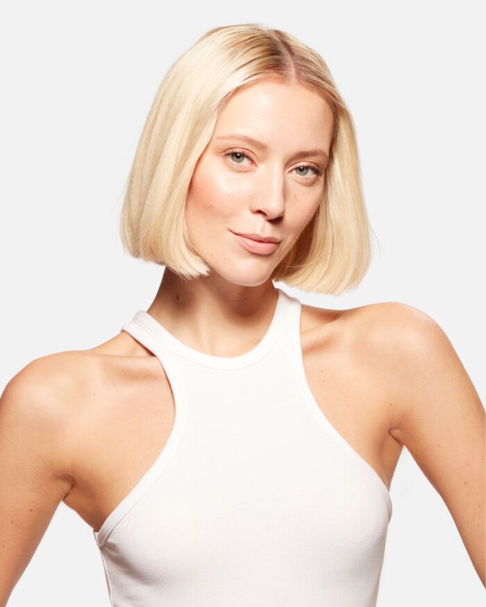 Une femme aux cheveux blonds courts, portant un debardeur blanc, regarde la camera avec un leger sourire.