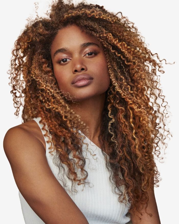 Une femme au teint caramel avec de longs cheveux boucles et ondules, regardant directement l'objectif avec une expression confiante. Elle porte un haut blanc sans manches.