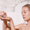 A person with wet hair holds a small jar in one hand, looking thoughtfully at the contents, in a marble bathroom setting.