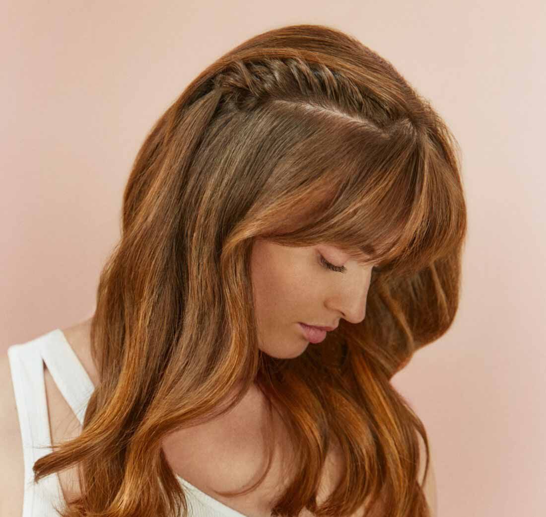 A woman with long, wavy, chestnut hair featuring a side braid and bangs, looking down against a soft pink background.