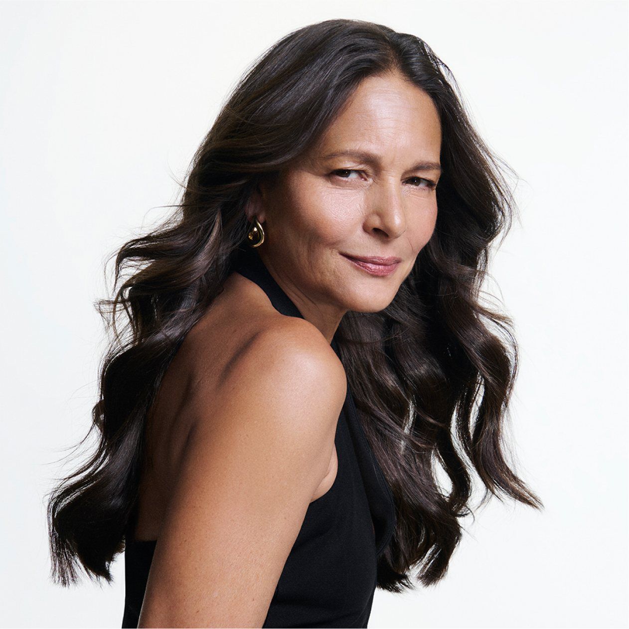 Portrait d’une femme mure aux longs cheveux sombres ondules avec un sourire assure sur fond blanc.