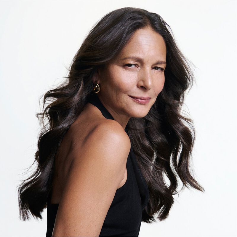 Portrait d’une femme mure aux longs cheveux sombres ondules avec un sourire assure sur fond blanc.