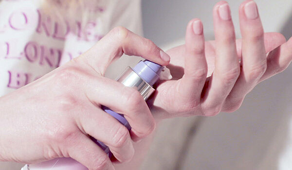 A close-up of a person's hand dispensing a small amount of cream from a lavender-colored pump bottle onto their palm.