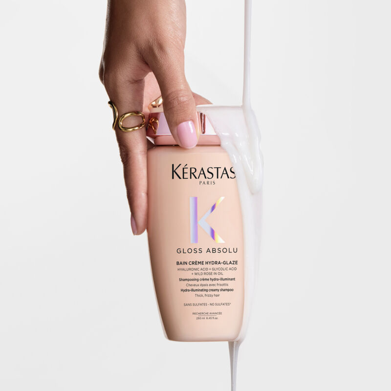 Main tenant un flacon de shampooing Kerastase Gloss Absolu avec un liquide blanc cremeux qui coule dessus.