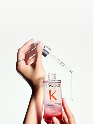 Gros plan sur des mains tenant une bouteille de Kerastase Genesis Serum Fortifiant, avec une pipette d'ou s'apprete a tomber une goutte de produit