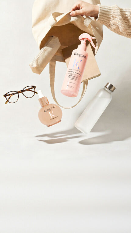 Trois flacons de produits capillaires Kerastase (Elixir Ultime, Genesis et Nutritive) sont disposes sur une surface blanche a cote d'un appareil photo vintage et de plusieurs pivoines blanches aux feuilles vertes. Le flacon dore Elixir Ultime est a gauche avec une pompe noire, le flacon rose Genesis avec un compte-gouttes argente est au centre, et le flacon degrade rouge-orange Nutritive avec un compte-gouttes argente est a droite. L'appareil photo argente avec sa dragonne noire se trouve a gauche des produits.